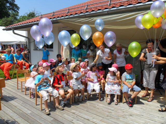Kinder sitzen auf Bänken und schauen erfreut zu bunten Luftballons, die von Erwachsenen gehalten werden.