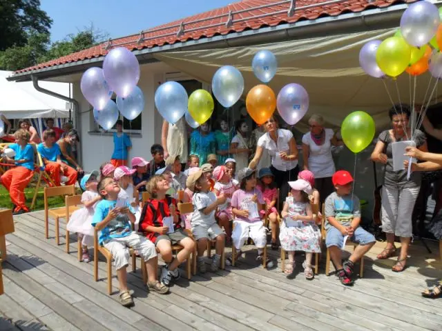 Kinder sitzen auf Bänken und schauen erfreut zu bunten Luftballons, die von Erwachsenen gehalten werden.